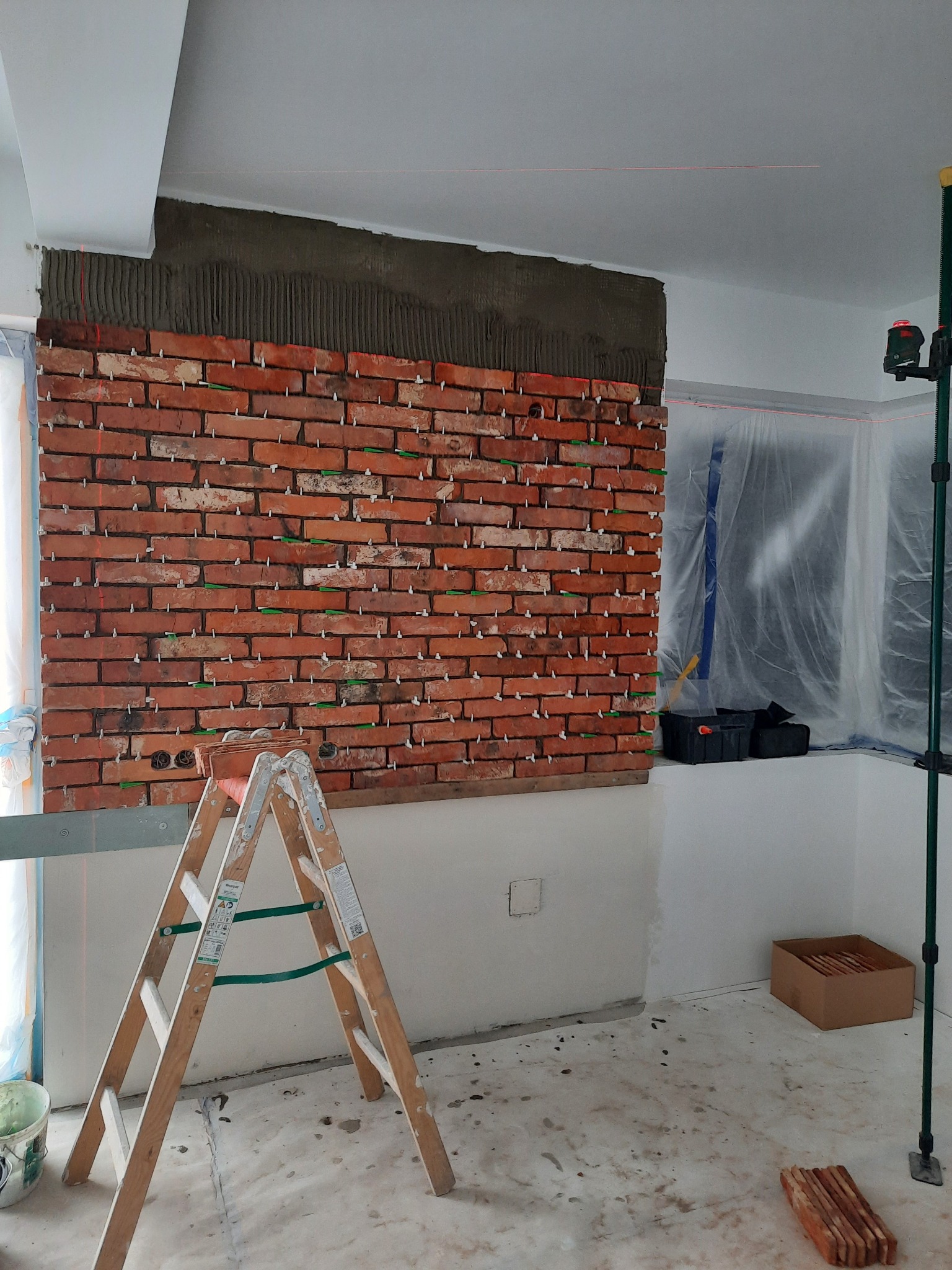 Montaż cegieł dekoracyjnych na ścianie, widoczny fragment ściany z przyklejonymi cegłami, szpachelka z zaprawą nad cegłami, poziomica laserowa i drabina na podłodze.
