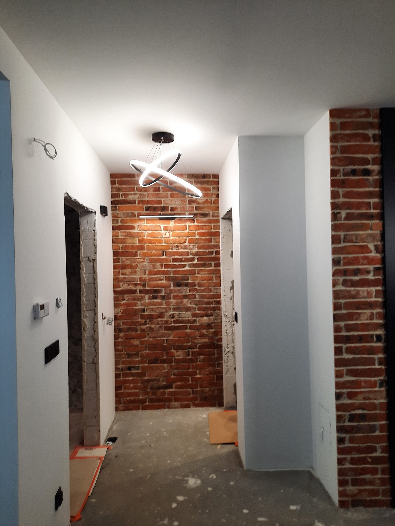 Wnętrze w trakcie remontu: ściana z czerwonej cegły, nowoczesna lampa sufitowa, surowe wykończenie przejść między pomieszczeniami, zabezpieczona podłoga.