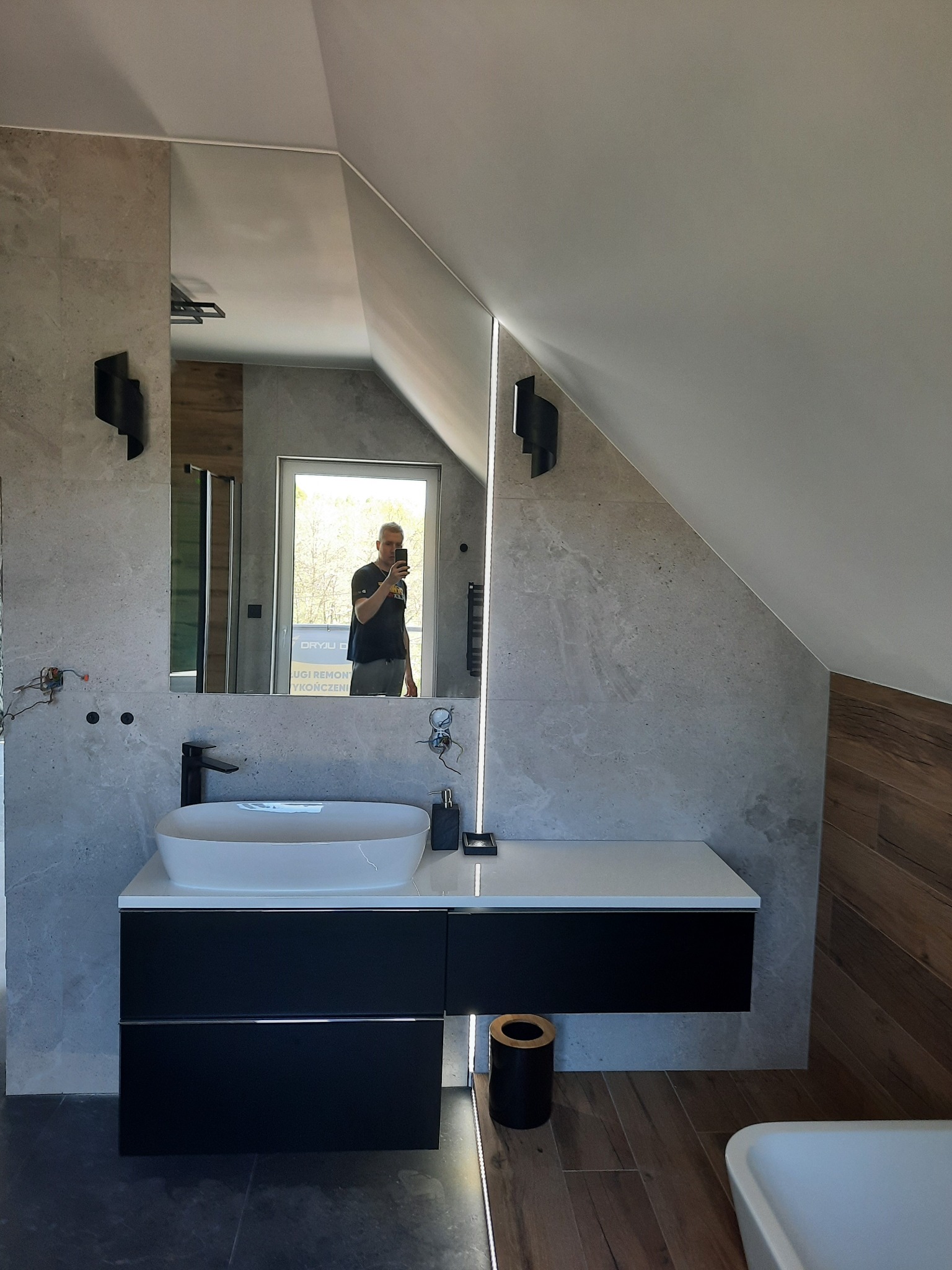 Nowoczesna łazienka na poddaszu z umywalką nablatową, czarną szafką pod umywalką, szarą płytką imitującą kamień, oświetleniem LED i lustrem, w którym odbija się osoba robiąca zdjęcie.
