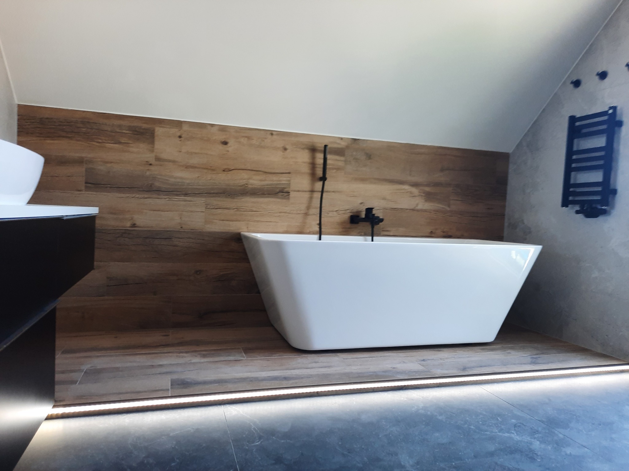 Nowoczesna łazienka na poddaszu z wolnostojącą białą wanną, drewnopodobną okładziną ściany i podświetleniem LED przy podłodze.