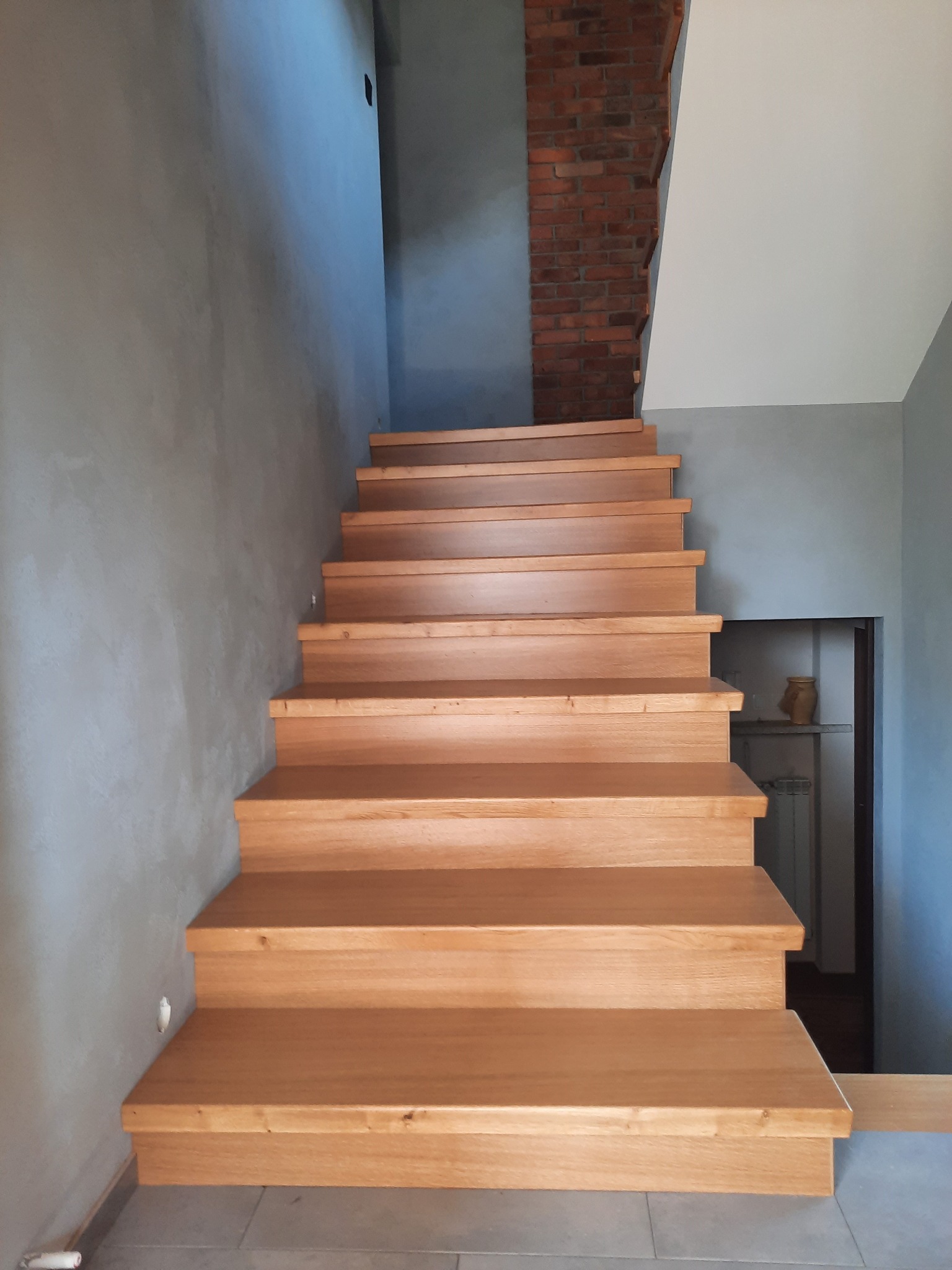 Drewniane schody z dębowymi stopniami na betonowej posadzce, ściana z cegły w tle, szare ściany, minimalistyczny design.