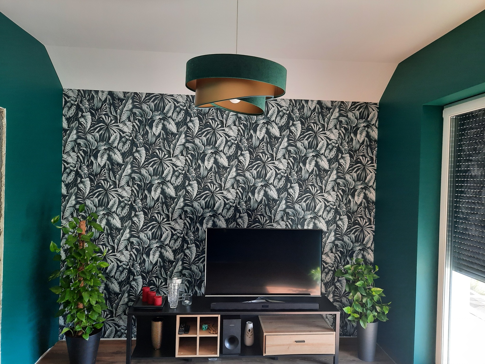 Pokój dzienny z akcentem na szmaragdowozielone ściany i tapetę w tropikalne liście, oświetlony nowoczesną, dwuwarstwową lampą wiszącą w odcieniach zieleni i złota, z roślinami doniczkowymi po...