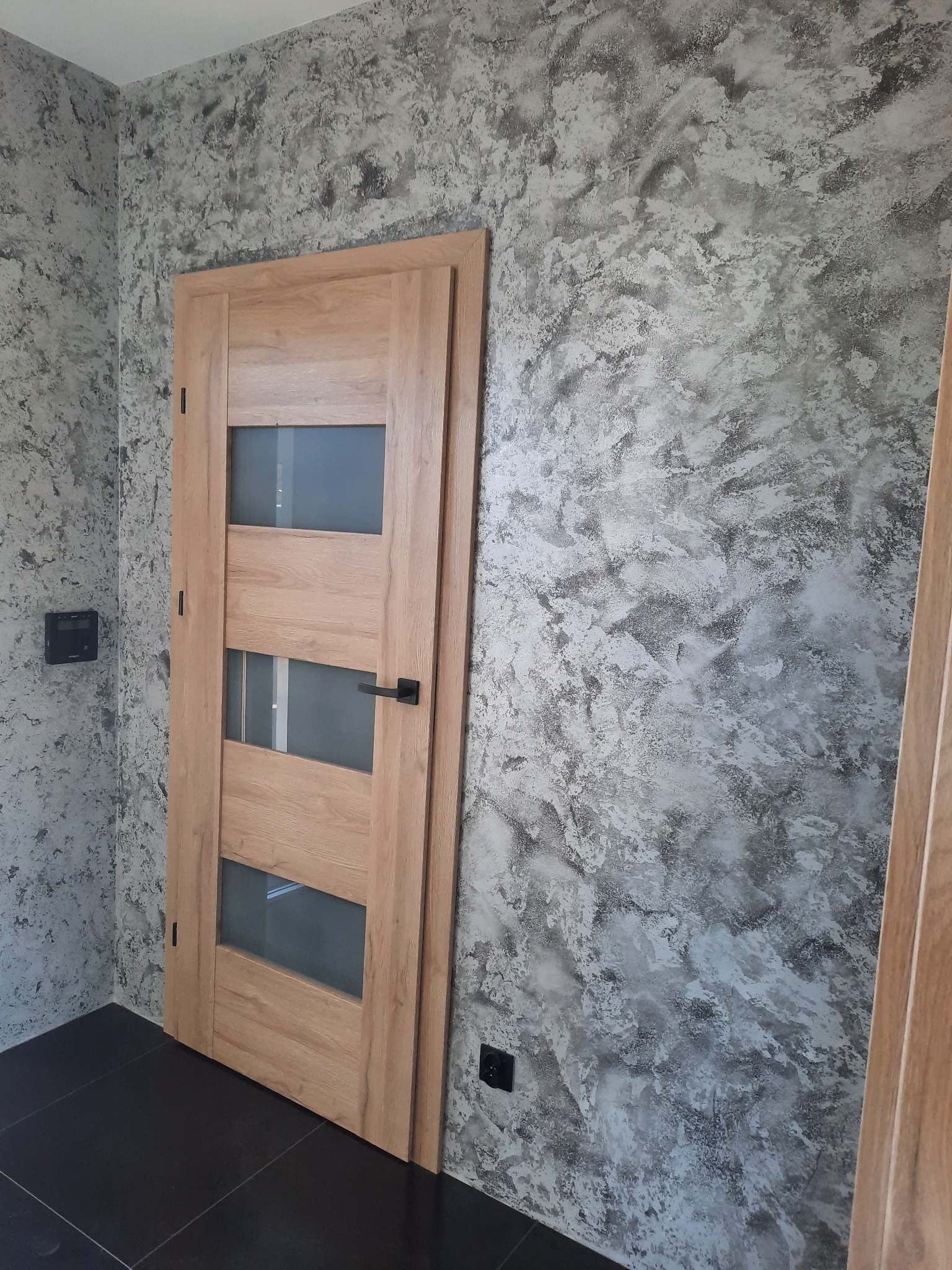 Drzwi wewnętrzne z przeszkleniami w drewnianej okleinie, zamontowane w pomieszczeniu z dekoracyjną, szaro-białą fakturą na ścianach i czarną, błyszczącą podłogą.