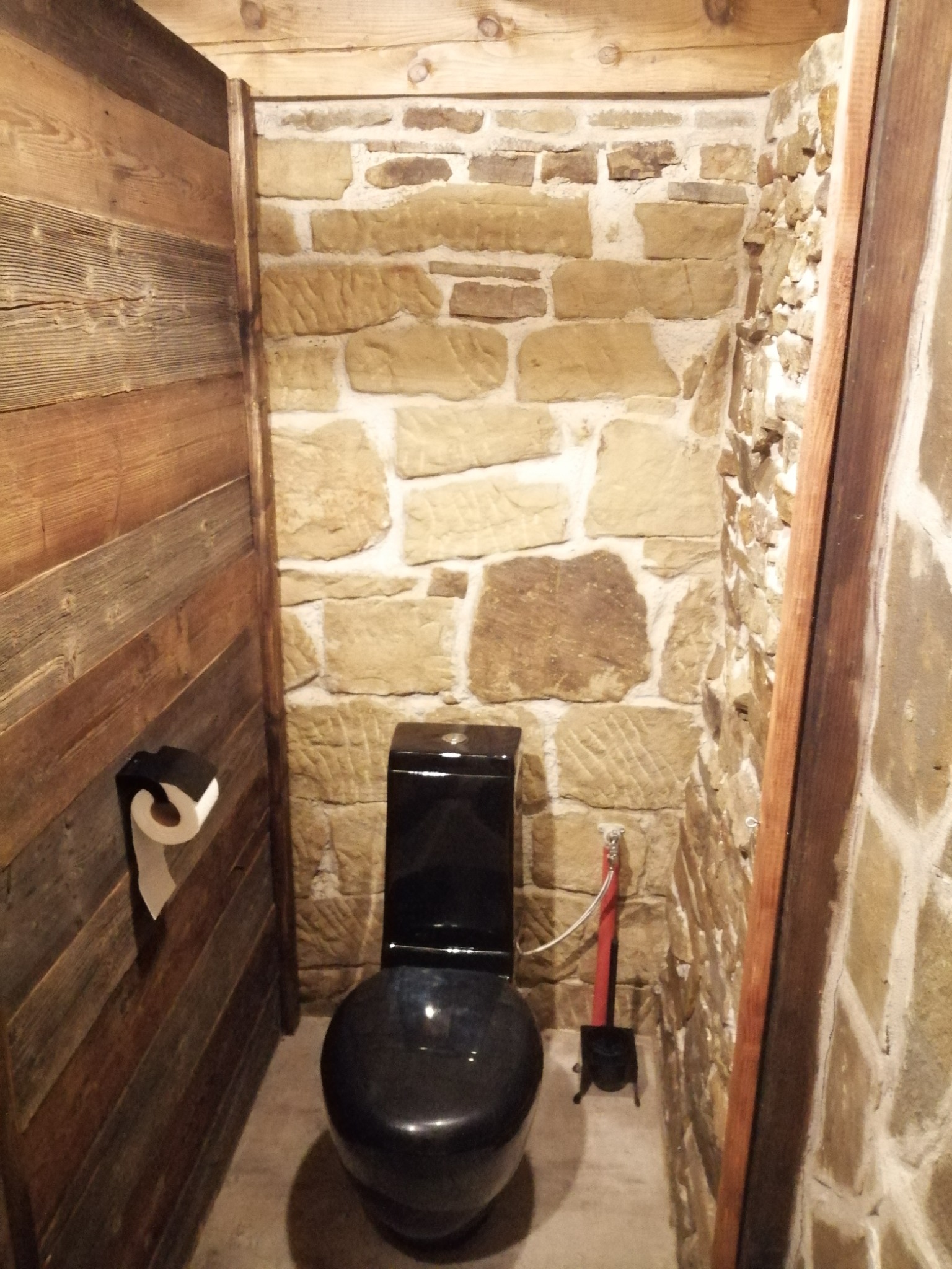Mała łazienka z czarną toaletą, ściany wykończone kamieniem i drewnem, widoczny podajnik na papier toaletowy i szczotka do toalety.