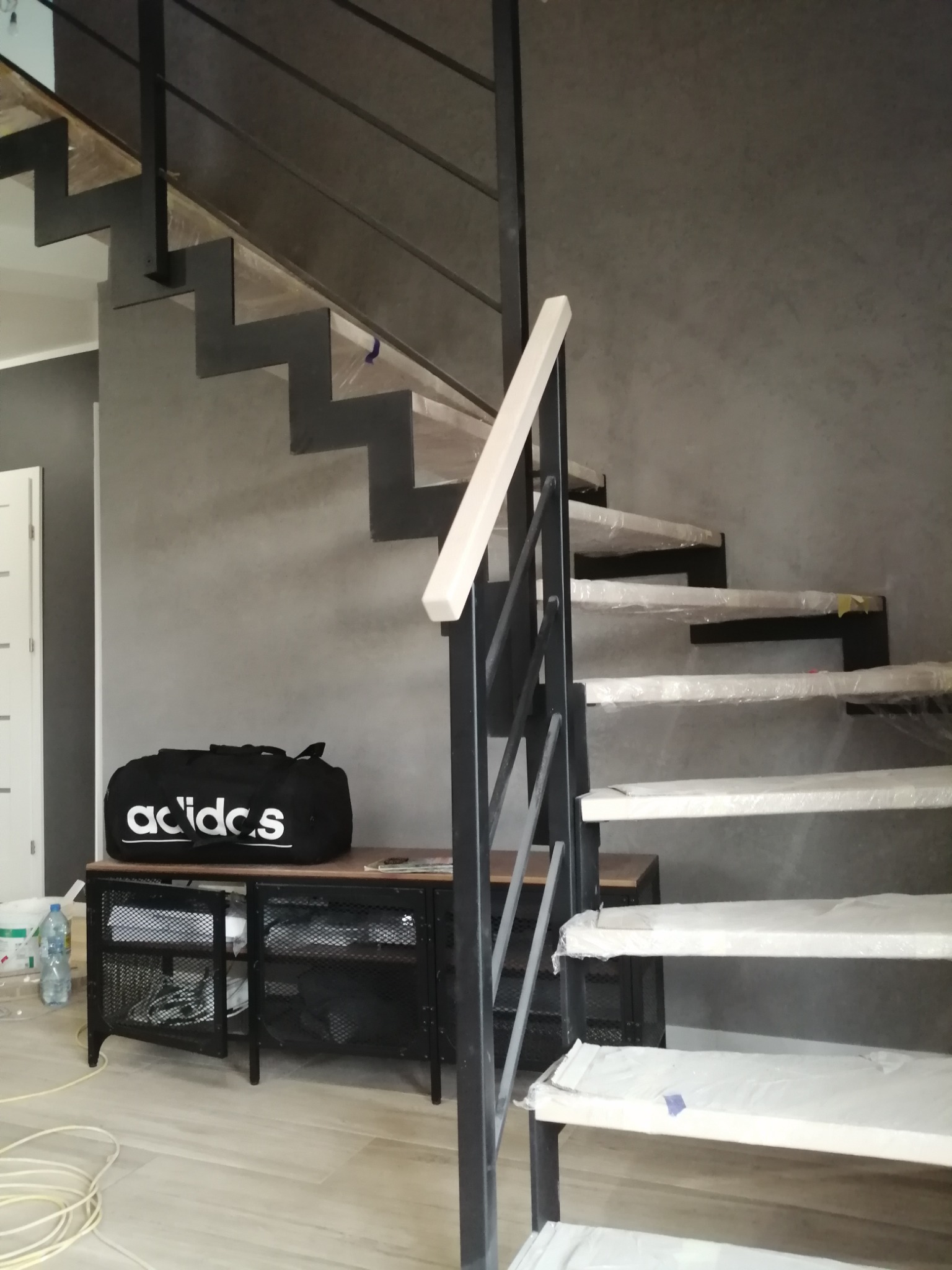 Nowoczesne schody stalowe z drewnianymi stopniami, zabezpieczone folią ochronną, w surowym wnętrzu z szarą ścianą i czarną szafką z torbą sportową Adidas