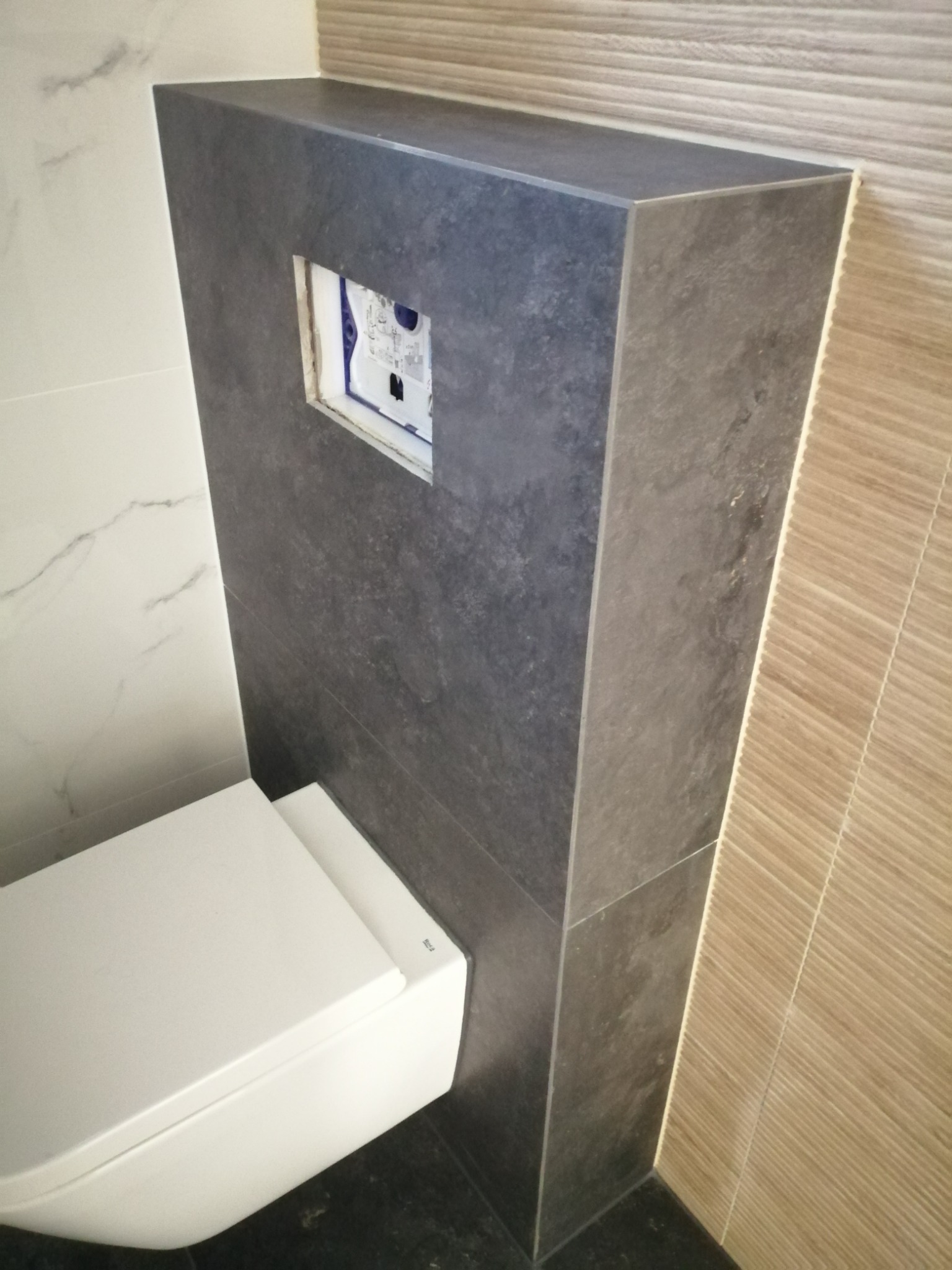 Podwieszana toaleta z zabudową z ciemnych, matowych płytek imitujących kamień, z otworem na panel sterujący, zamontowana na tle białych i beżowych płytek w łazience.