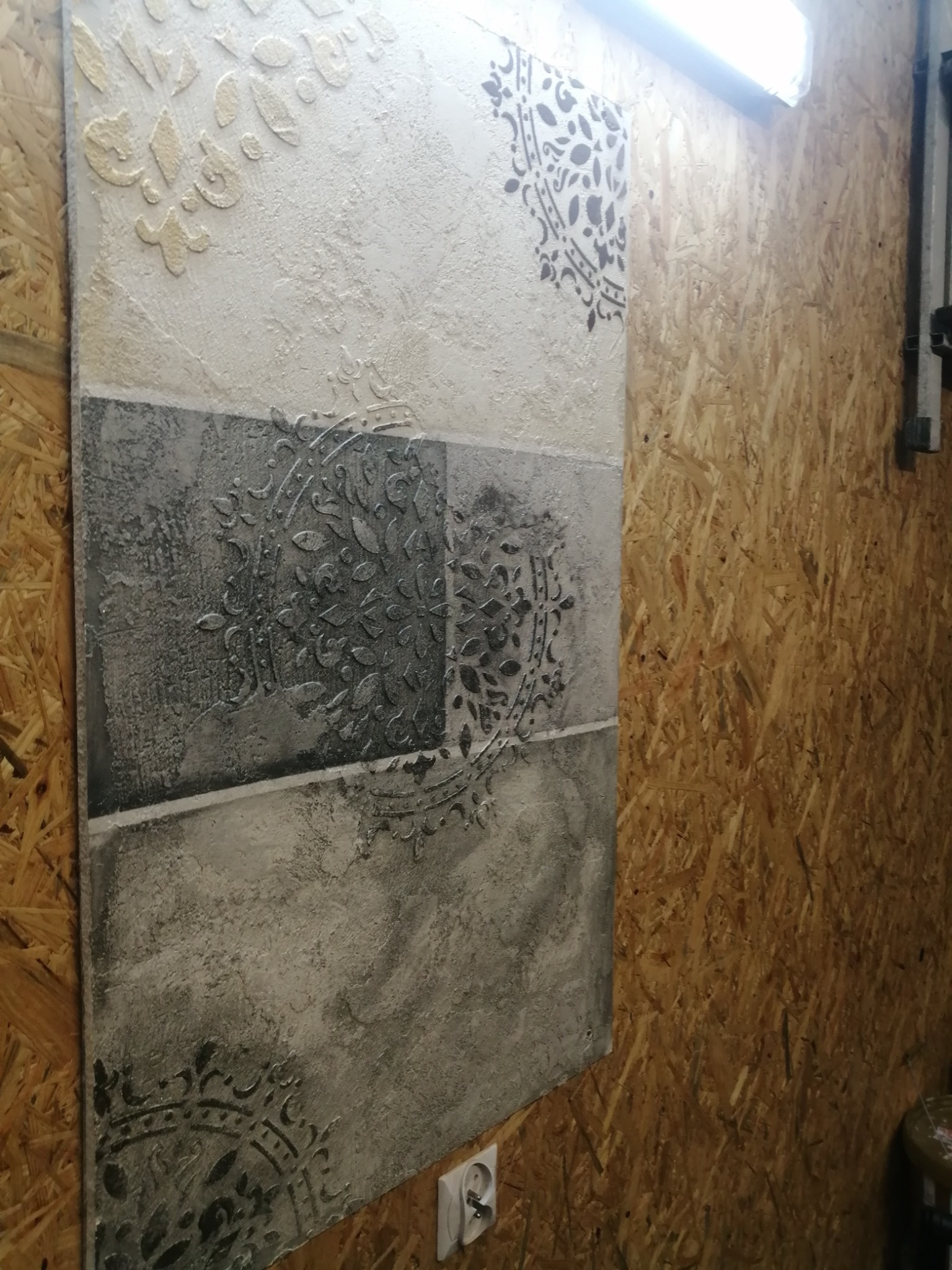 Panel dekoracyjny z efektem strukturalnym, podzielony na poziome pasy z różnymi wzorami i fakturami, umieszczony na ścianie z płyty OSB, oświetlony lampą jarzeniową.