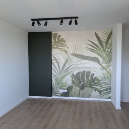 PONTSIR DOROTA PONTSIR - Wykończone wnętrze pokoju z drewnianą podłogą, ścianą z motywem roślinnym i czarnym panelem, oświetlone reflektorami.