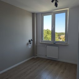 PONTSIR DOROTA PONTSIR - Wnętrze pokoju po remoncie: drewniana podłoga, szare ściany, białe okno z widokiem na zieleń, nowoczesne oświetlenie sufitowe i ścienne. Grzejnik pod oknem.