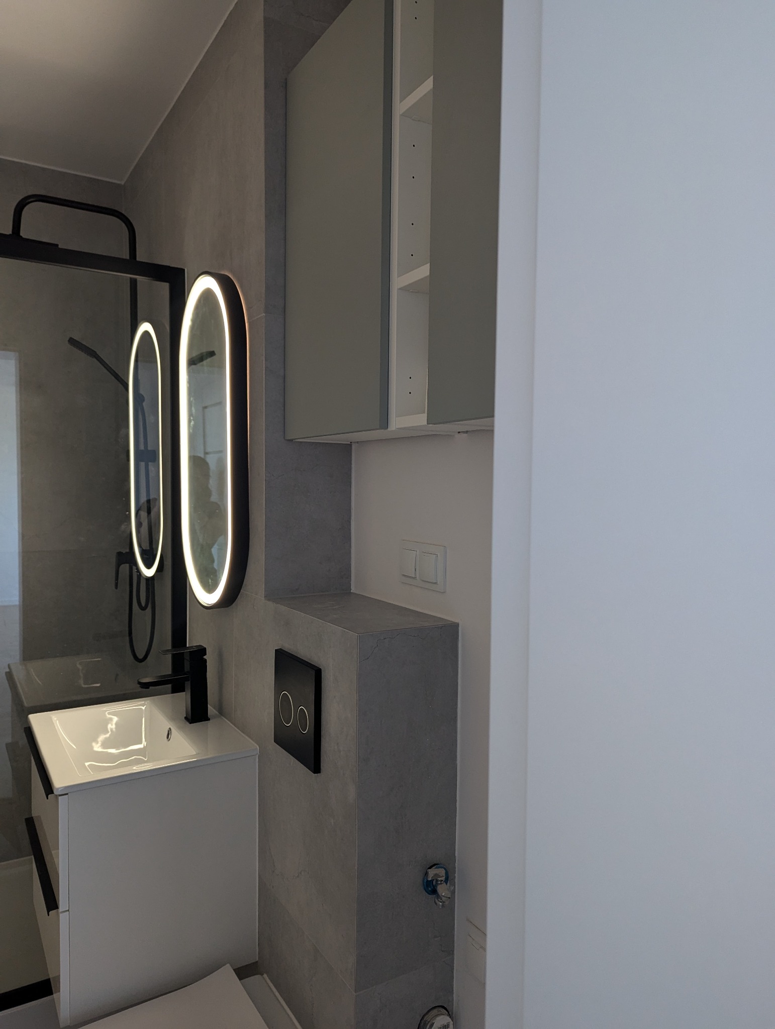 Nowoczesna łazienka po remoncie: szara płytka, podświetlane lustra owalne, biała szafka z umywalką i czarna bateria. Minimalistyczny design, widok na kabinę prysznicową.