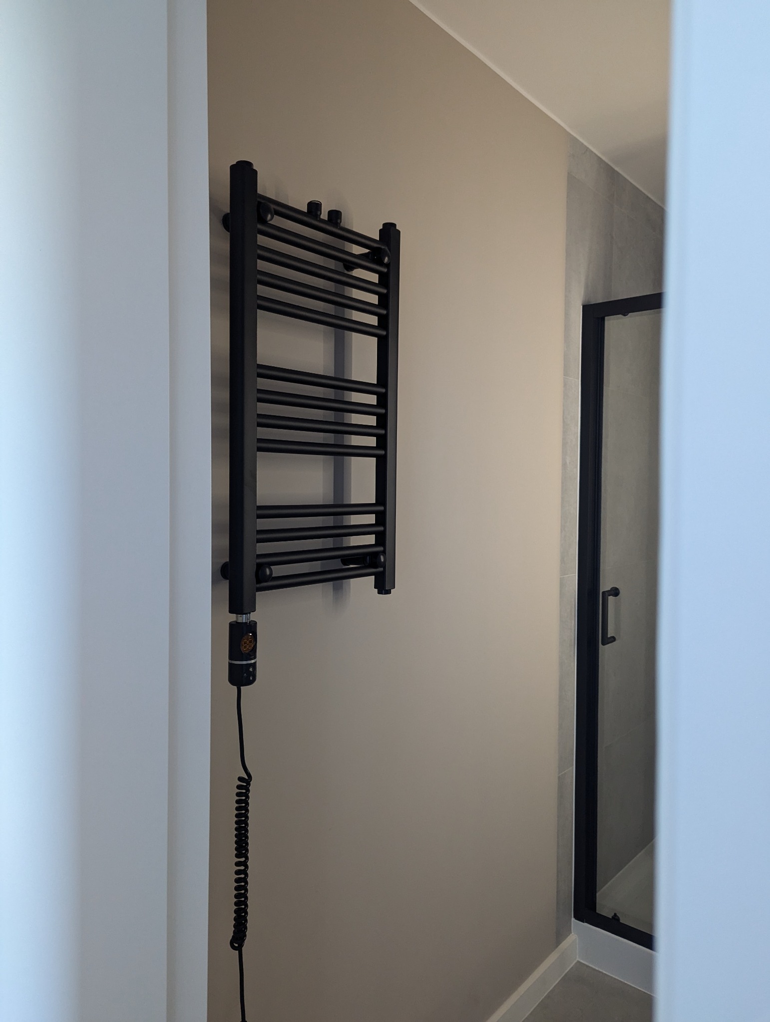 Czarny, elektryczny grzejnik łazienkowy na beżowej ścianie, widoczny fragment kabiny prysznicowej z czarną ramą. Minimalistyczny design, nowoczesna łazienka.