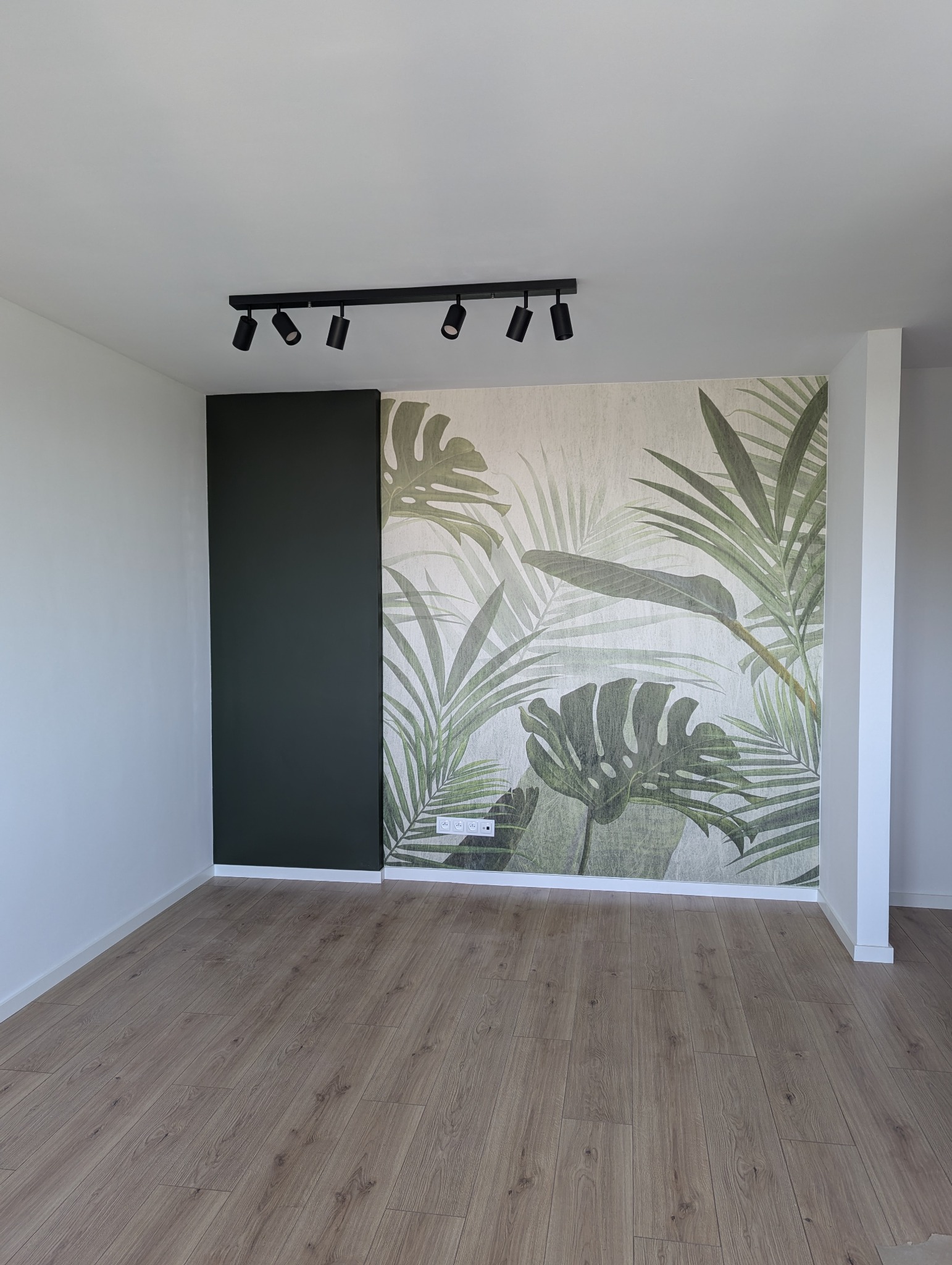 Wykończone wnętrze pokoju z drewnianą podłogą, ścianą z motywem roślinnym i czarnym panelem, oświetlone reflektorami.
