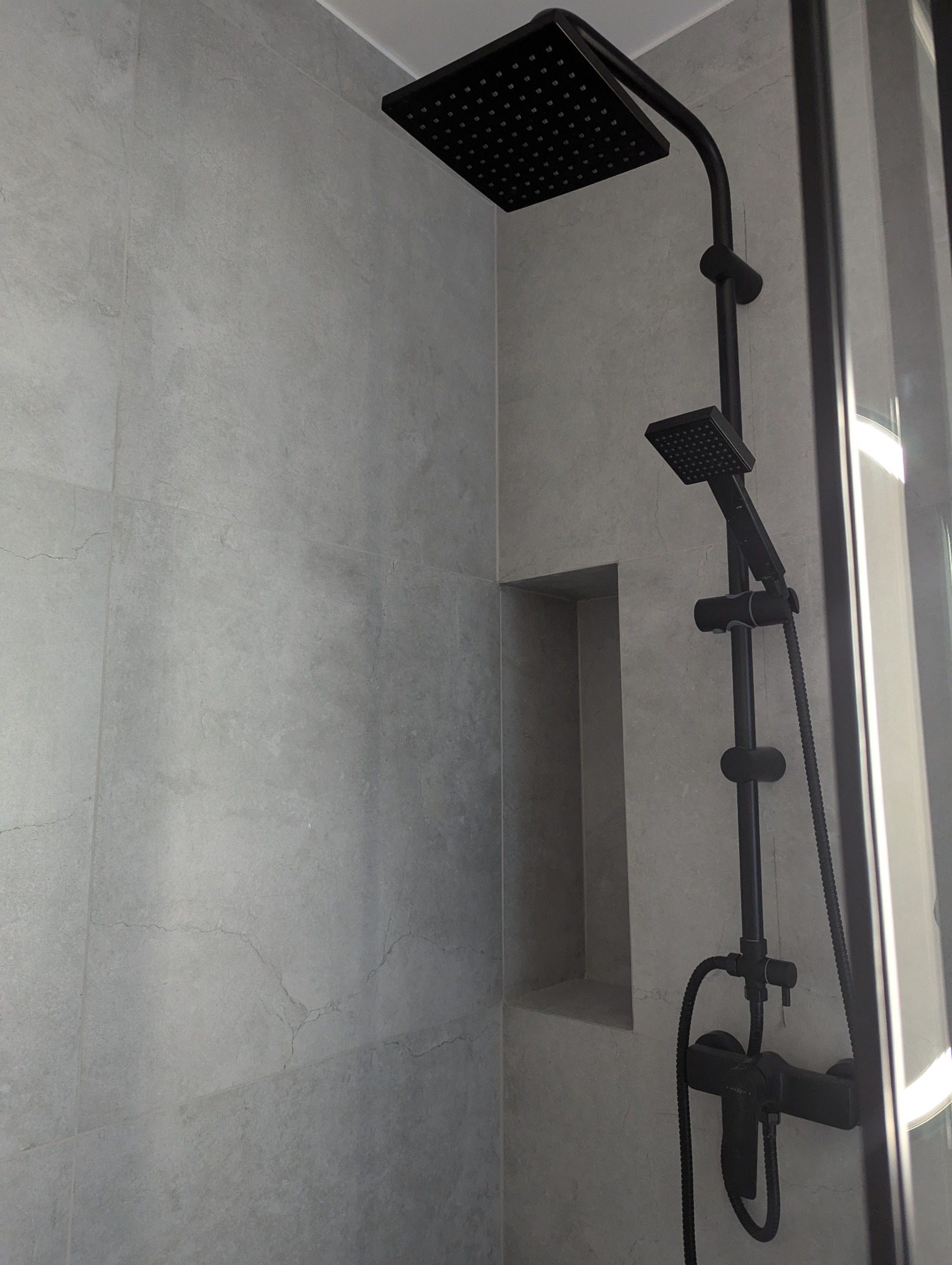 Nowoczesna kabina prysznicowa z czarną armaturą i wnęką w szarej, imitującej kamień, płytce. Minimalistyczny design z wyraźnym kontrastem kolorystycznym.