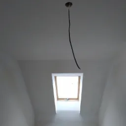 Poddasze w trakcie wykańczania: widoczne okno dachowe, kabel zwisający z sufitu, ślady szpachlowania na ścianach i drewniana listwa na dole ściany.