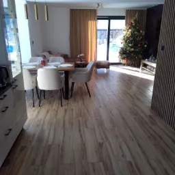 Nowoczesne wnętrze w Żywcu z drewnopodobną podłogą, jadalnią z białymi krzesłami i świąteczną choinką w tle, widok z perspektywy wejścia.