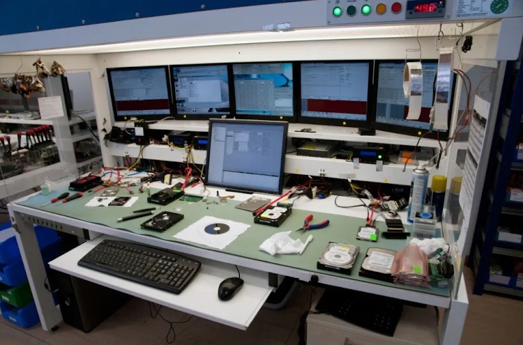 Stanowisko pracy z pięcioma monitorami, klawiaturą, myszką i rozłożonymi dyskami twardymi oraz narzędziami na stole, w tle widoczne półki z akcesoriami.