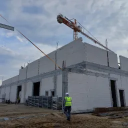 Budowa hali z białych bloczków betonu komórkowego, widoczny żuraw budowlany, pracownik w kamizelce odblaskowej idący po piaszczystym terenie.