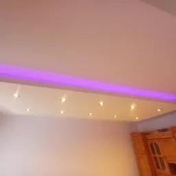 Podwieszany sufit z oświetleniem punktowym i fioletową listwą LED, widoczny fragment drewnianej szafy w rogu pokoju.