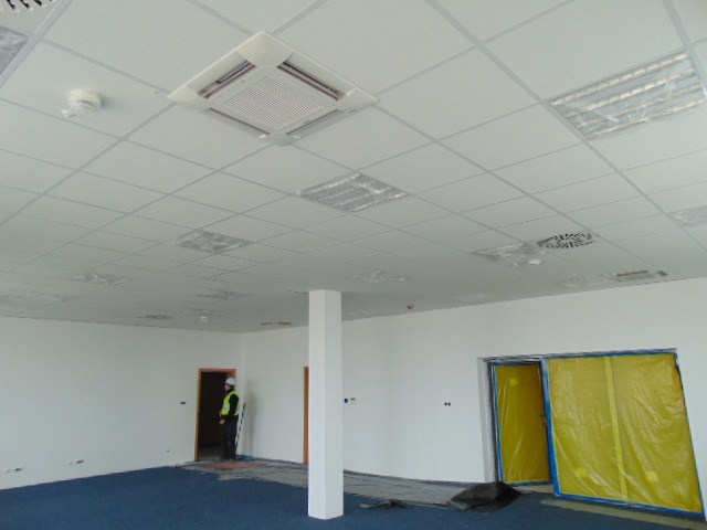 Montaż sufitu podwieszanego z kratką wentylacyjną i oświetleniem w biurze, widoczny pracownik w kamizelce odblaskowej i zabezpieczone folią drzwi.