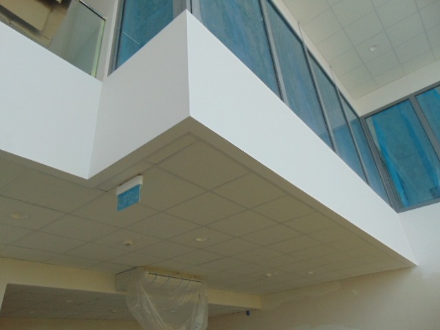 Sufit podwieszany kasetonowy z widoczną instalacją klimatyzacyjną zabezpieczoną folią, oświetlenie punktowe i fragment ściany z oknami w nowoczesnym biurze.