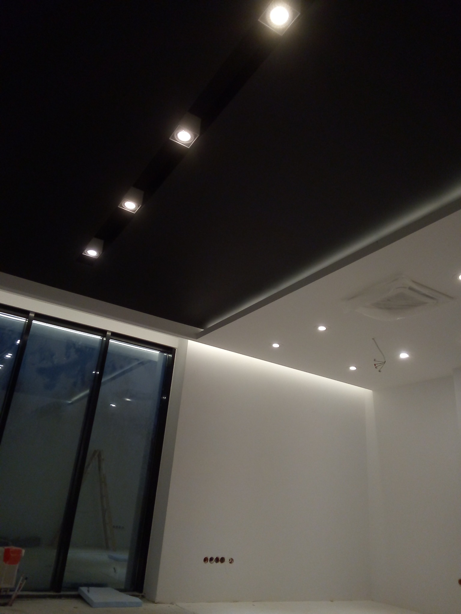 Częściowo wykończone wnętrze z czarnym, podwieszanym sufitem z punktowym oświetleniem, białe ściany z widocznymi gniazdkami elektrycznymi i dużym oknem, widoczne elementy instalacji klimatyzacji...