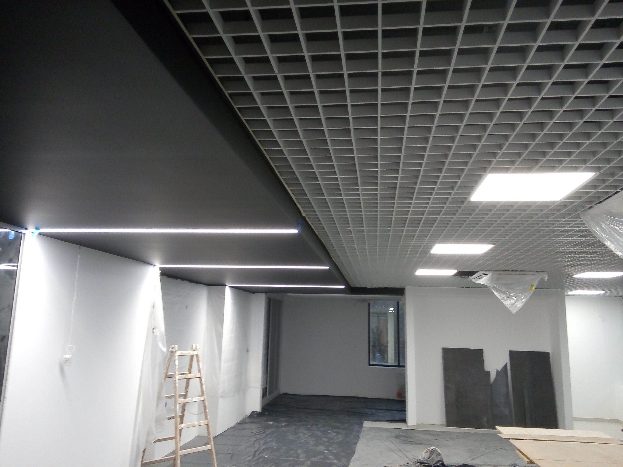 Montaż sufitu podwieszanego z rastrem i oświetleniem liniowym w biurze, widoczna drabina malarska i zabezpieczona folią podłoga.