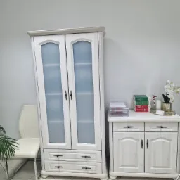 Jasne biuro: biała szafa z przeszklonymi drzwiami, komoda z dokumentami i książkami prawniczymi, białe krzesło, orchidea w złotym wazonie.