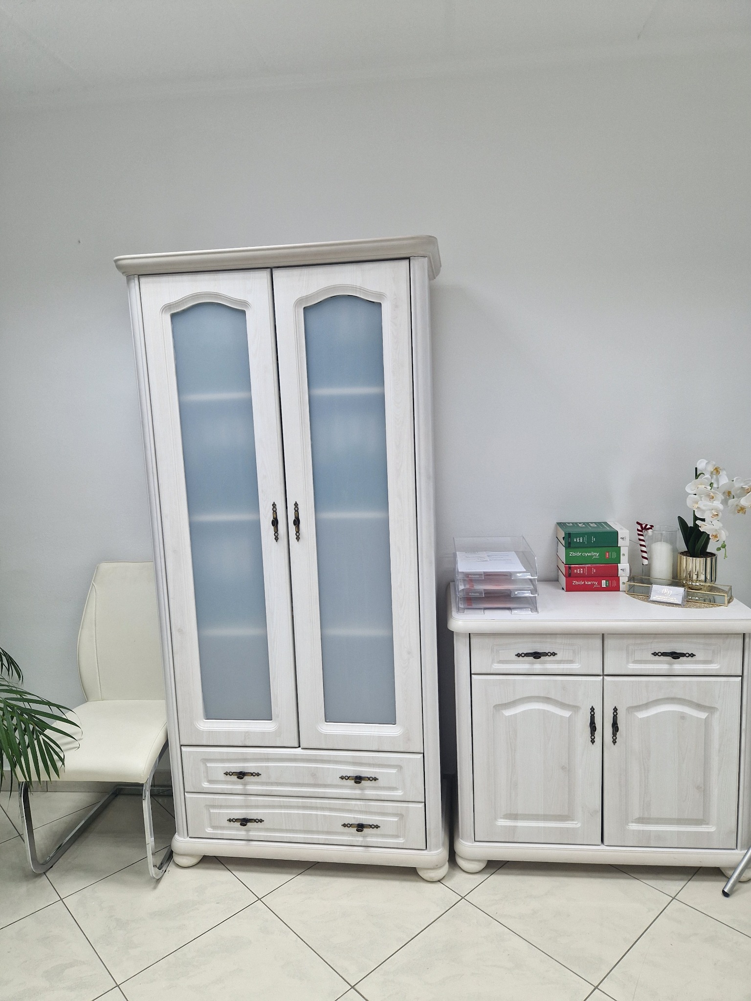 Jasne biuro: biała szafa z przeszklonymi drzwiami, komoda z dokumentami i książkami prawniczymi, białe krzesło, orchidea w złotym wazonie.