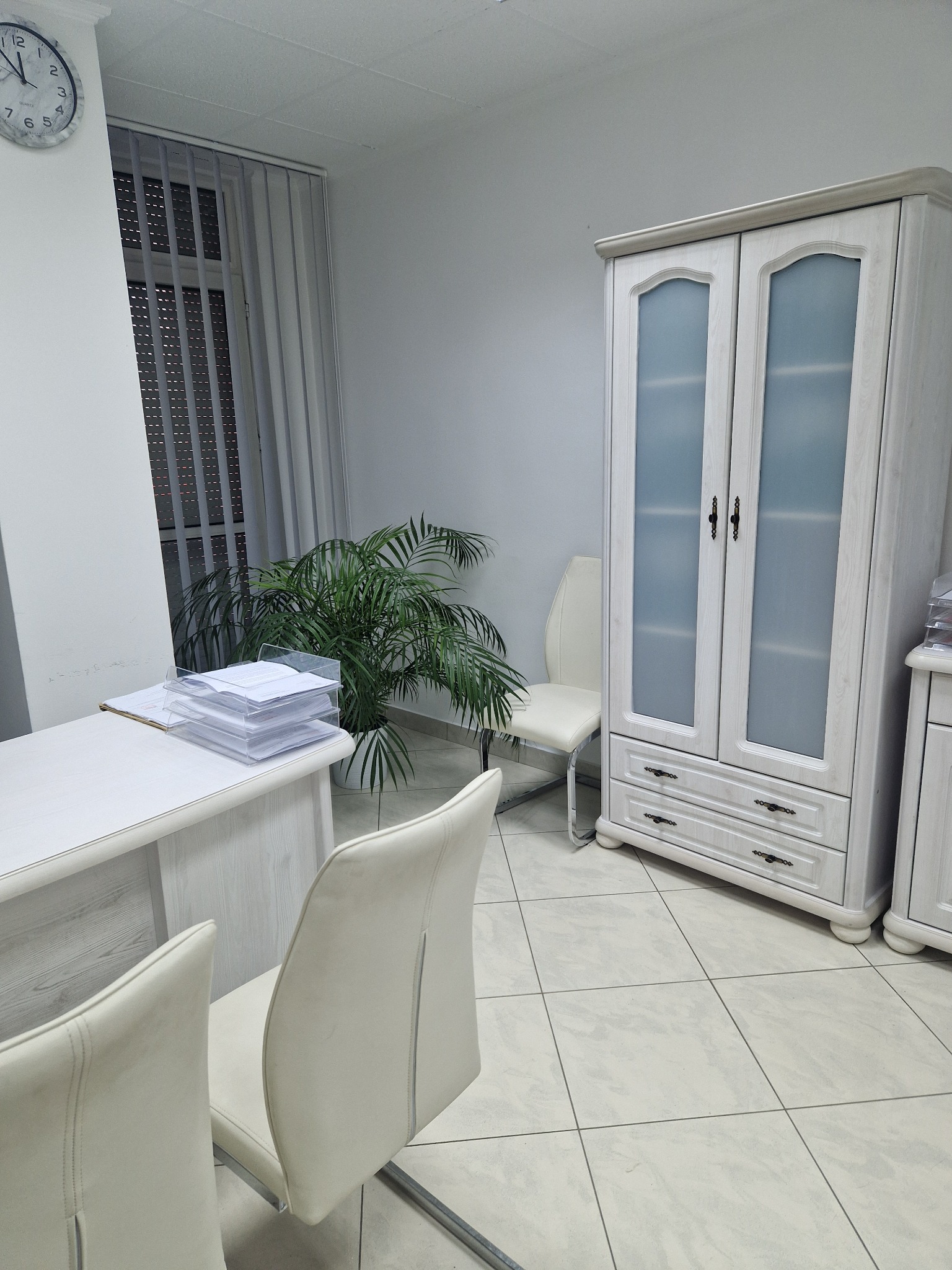 Jasne biuro z białymi meblami: biurko z dokumentami, szafa z przeszklonymi drzwiami, krzesła z białej skóry ekologicznej, zegar na ścianie i roślina doniczkowa.