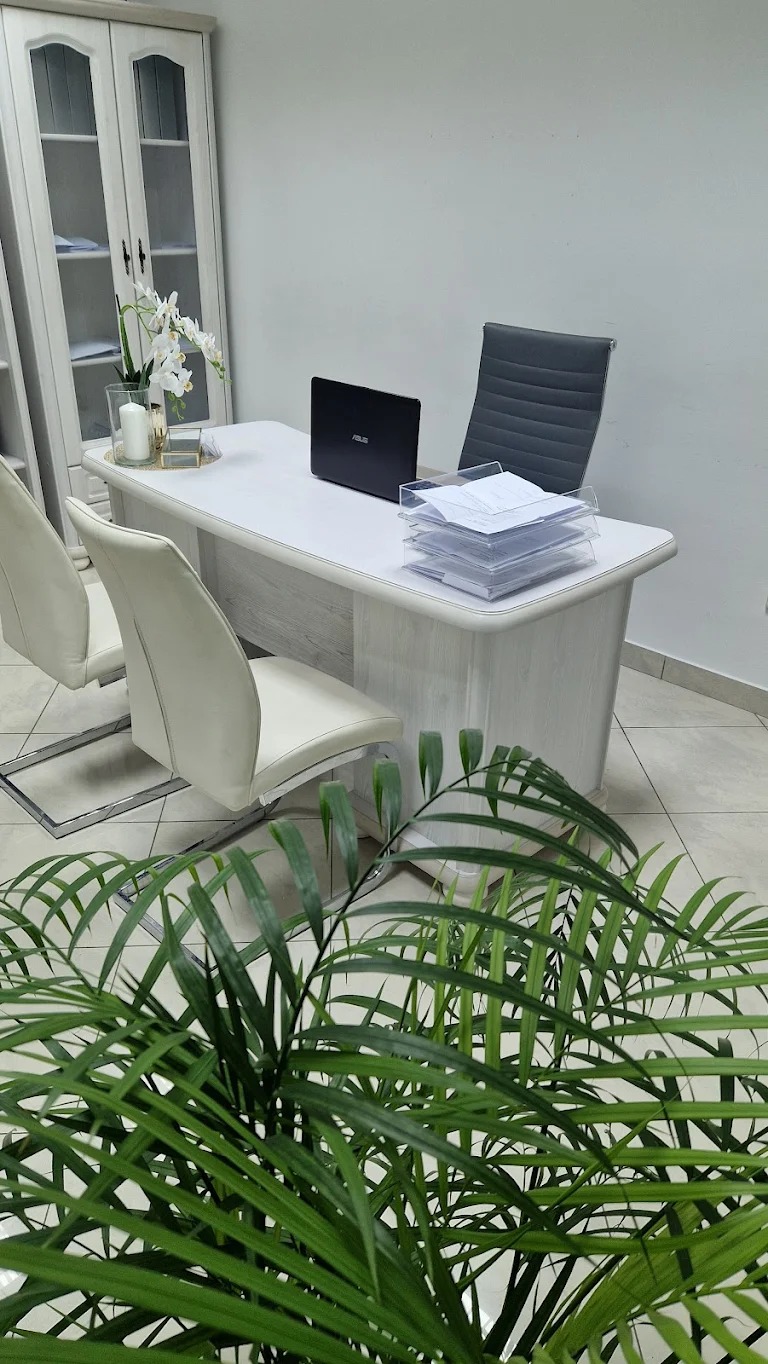 Jasne biuro z biurkiem, laptopem i dokumentami, oświetlone naturalnym światłem. Roślina w doniczce dodaje świeżości. Widoczny regał na dokumenty.