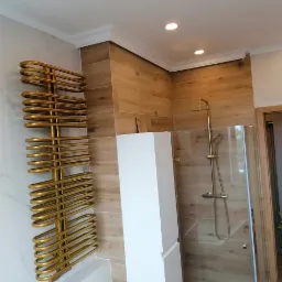 Nowoczesna łazienka z drewnopodobnymi płytkami, złotym grzejnikiem drabinkowym i kabiną prysznicową ze złotą armaturą. Widoczna biała szafka wisząca i oświetlenie punktowe.