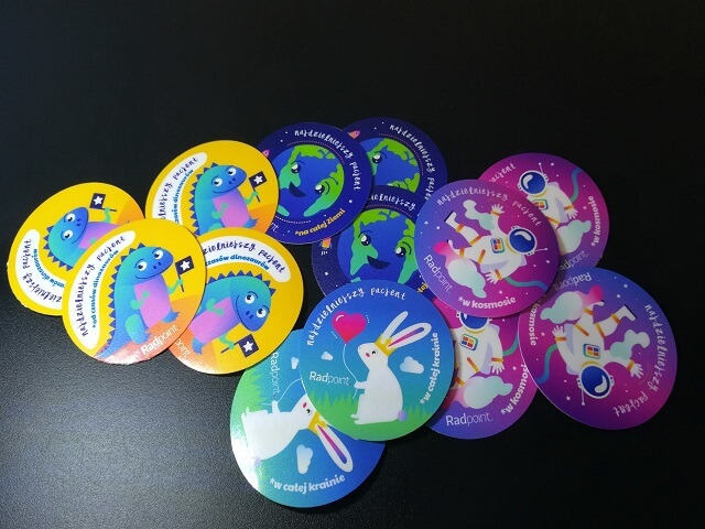 Kolekcja okrągłych, foliowych nalepek z różnymi motywami: dinozaury, planeta Ziemia z uśmiechem, kosmonauta i królik z balonem, każda z logo 'Radpoint' i napisem 'najdzielniejszy pacjent'.