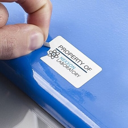 Biała, prostokątna etykieta z napisem 'PROPERTY OF NEXXON LABORATORY' i symbolem atomu, naklejana przez palec na niebieską powierzchnię. Etykieta posiada perforowaną zakładkę ułatwiającą odklejanie.