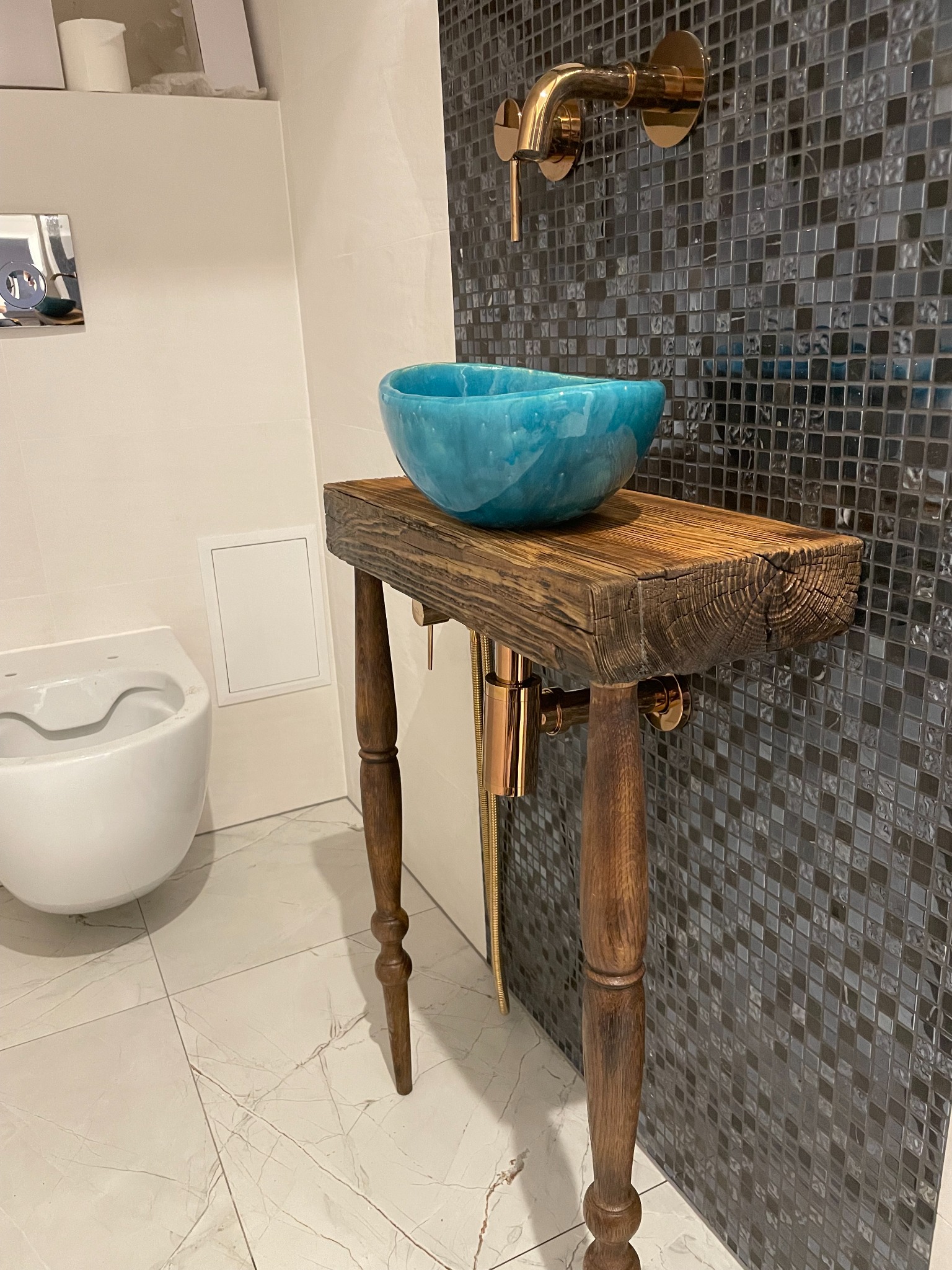 Turkusowa umywalka nablatowa na drewnianym blacie z toczonymi nogami, złota bateria na ścianie z mozaiki w odcieniach szarości, obok wisząca toaleta.