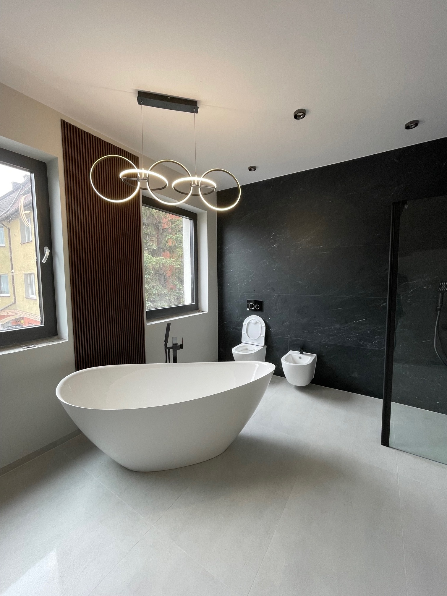 Nowoczesna łazienka z wolnostojącą białą wanną, czarną ścianą z imitacji marmuru i designerską lampą pierścieniową, widok na zewnątrz przez okno.
