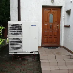 Biała jednostka zewnętrzna z logo Panasonic, umieszczona na czerwonym stelażu, stoi przed wejściem do domu z drewnianymi drzwiami i schodami z płytek.