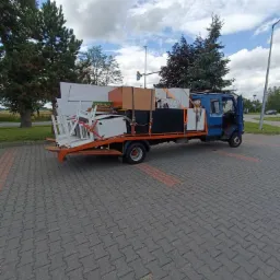 Niebieska laweta z otwartymi drzwiami kabiny, załadowana meblami i sprzętami, zabezpieczonymi pasami, na tle brukowanej powierzchni i zieleni.