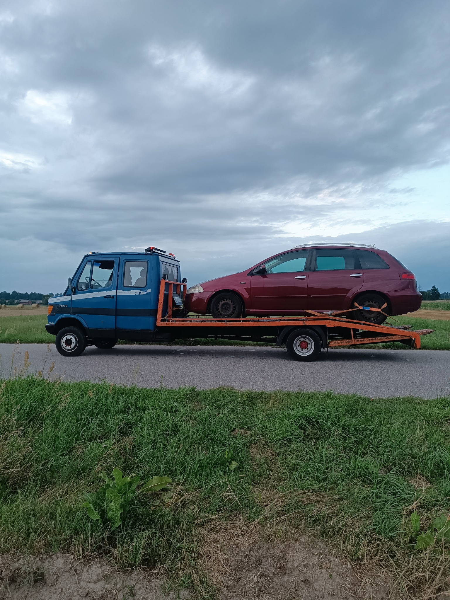 Niebieska laweta z czerwonym samochodem osobowym na platformie, zaparkowana na poboczu drogi, z polem w tle i pochmurnym niebem.
