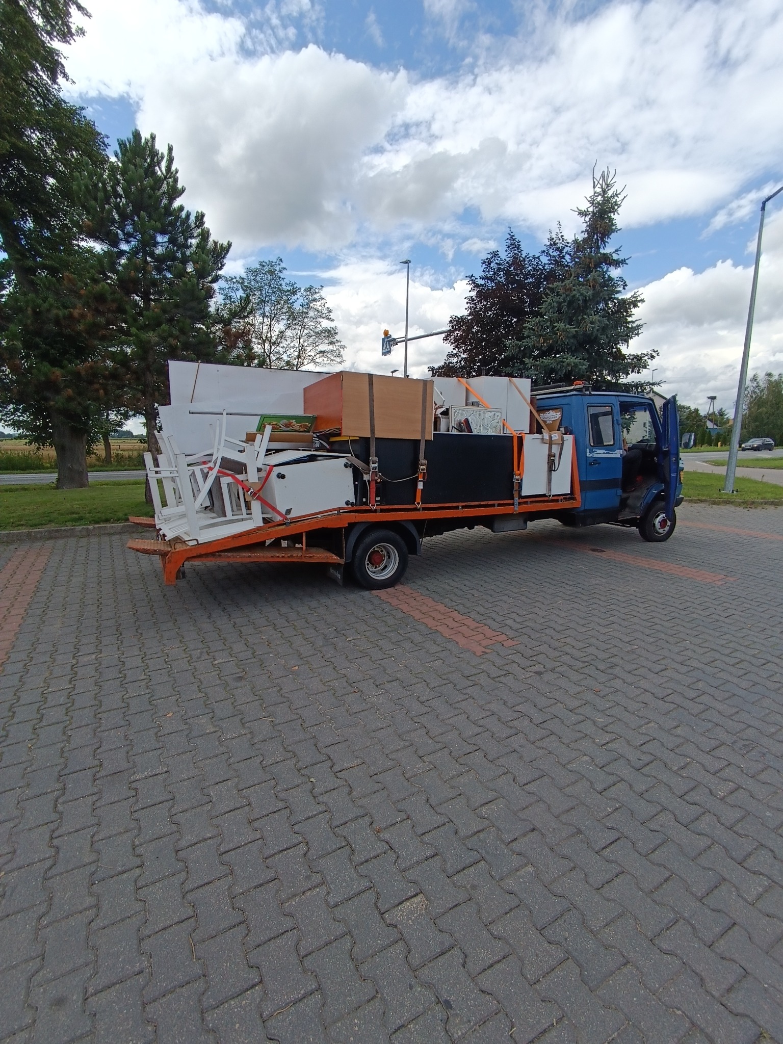 Niebieska laweta z otwartymi drzwiami kabiny, załadowana meblami i sprzętami, zabezpieczonymi pasami, na tle brukowanej powierzchni i zieleni.