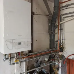 Nowy piec gazowy De Dietrich, jeszcze w folii ochronnej, zamontowany na ścianie z widocznymi rurami miedzianymi i plastikowymi odpływami kanalizacyjnymi, instalacja w trakcie podłączania.
