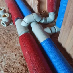 Fragment instalacji wodnej z rurami w izolacji czerwonej i niebieskiej, połączonymi kształtkami z tworzywa sztucznego, widoczne fragmenty muru i podłoża.