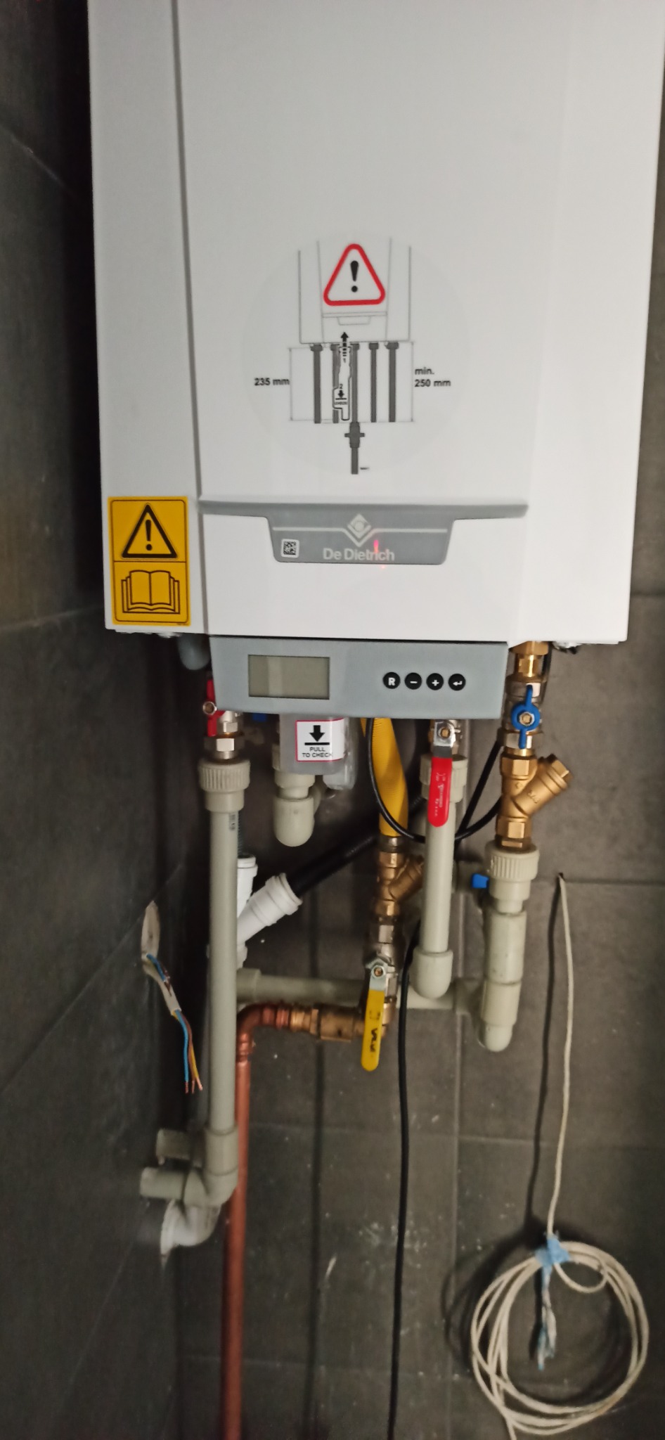 Zamontowany piec gazowy De Dietrich z widocznymi przyłączami hydraulicznymi i gazowymi, etykietą ostrzegawczą i schematem instalacji na obudowie, na tle ciemnej ściany z płytek.