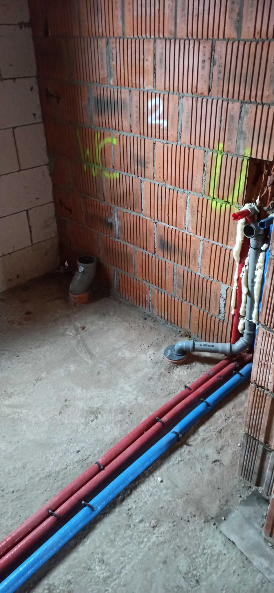 Surowy stan: Instalacja rur grzewczych i kanalizacyjnych w nowym budynku, widoczne czerwone i niebieskie rury, odpływ kanalizacyjny oraz fragmenty muru z pustaków ceramicznych i pianki montażowej.