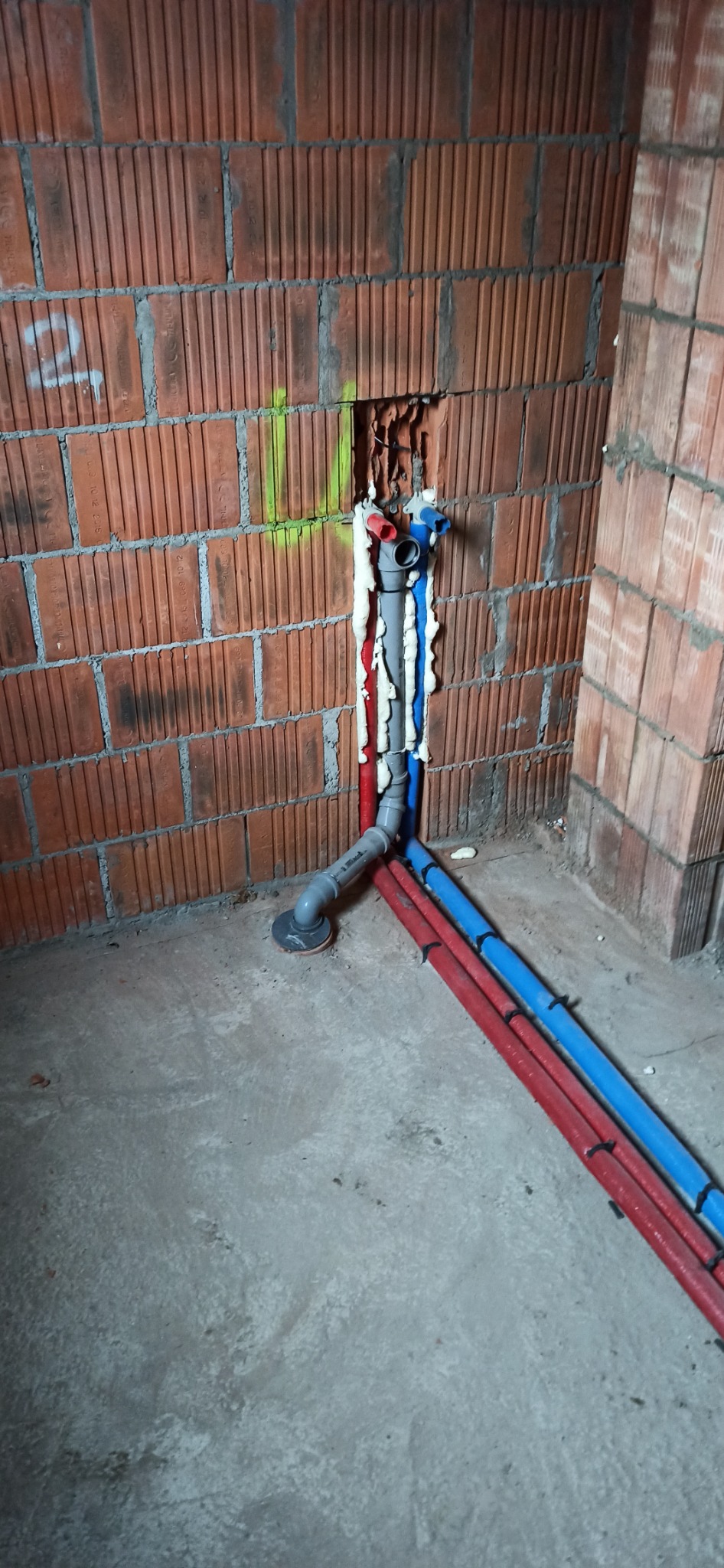 Wewnętrzne instalacje hydrauliczne w surowym stanie, rury PEX czerwone i niebieskie prowadzone wzdłuż ściany z cegły dziurawki, zabezpieczone pianką montażową w bruździe.