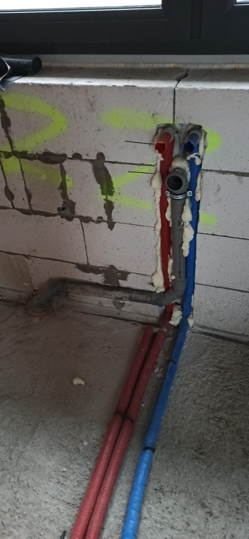 Czerwona i niebieska rura w ścianie z betonu komórkowego, uszczelnione pianką montażową, widoczny fragment szarej rury kanalizacyjnej.