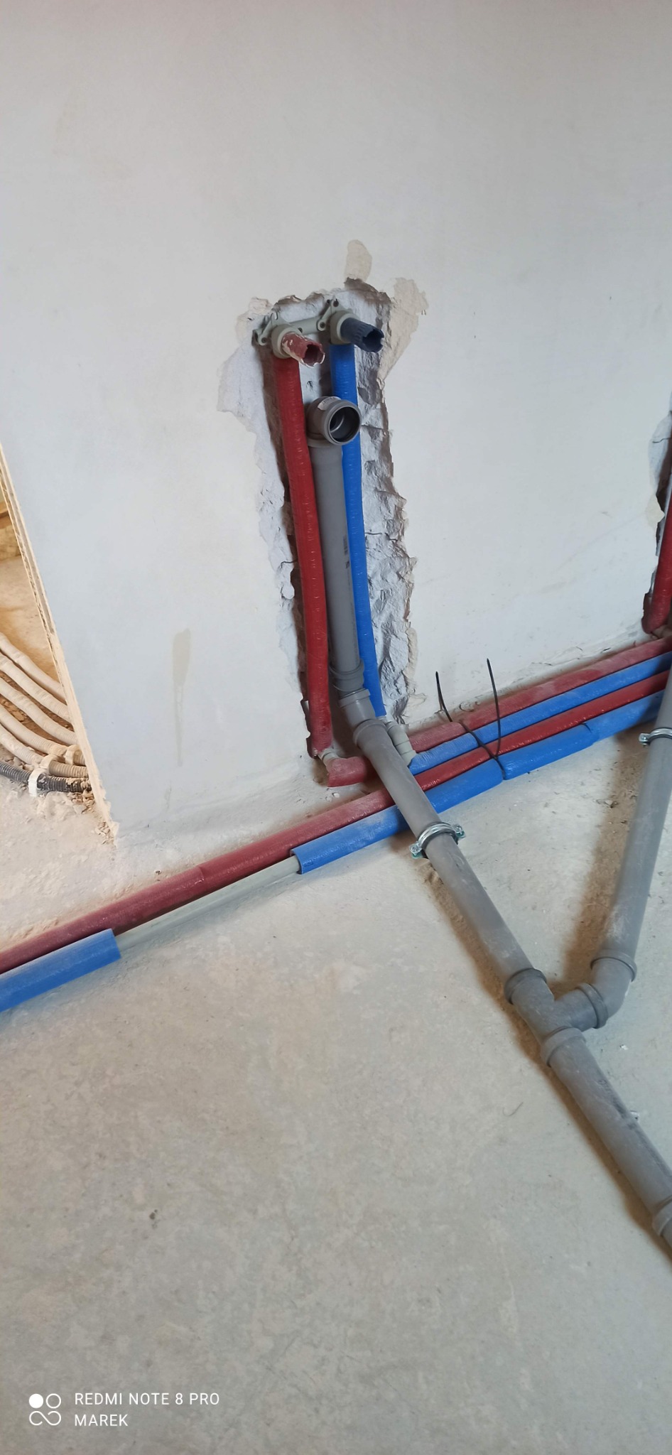 Wewnętrzna instalacja wodna w trakcie montażu: rury PEX w izolacji czerwonej i niebieskiej, szare rury kanalizacyjne z uchwytami mocującymi, widoczne połączenia i fragment ściany w stanie surowym.