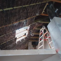 Poddasze w trakcie izolacji, widoczna wełna mineralna zabezpieczona sznurkami, fragment okna dachowego, drewniana drabina oparta o komin, surowy stan wykończenia.