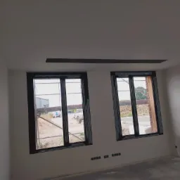 Wnętrze w trakcie wykończenia z dwoma oknami z brązowymi ramami, widoczne nieotynkowane ściany zewnętrzne i przygotowane otwory na gniazdka elektryczne.