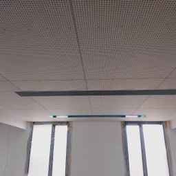 Usługi Budowlane Dawid Nabakowski - Sufit podwieszany z perforowanej płyty w pomieszczeniu w trakcie wykańczania, widoczne okna zabezpieczone folią.