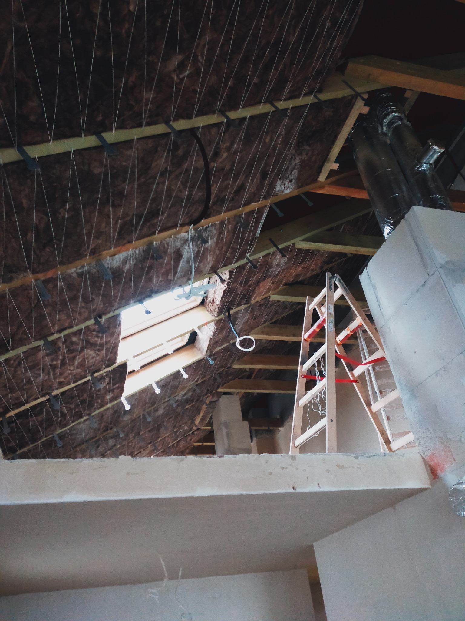 Poddasze w trakcie izolacji, widoczna wełna mineralna zabezpieczona sznurkami, fragment okna dachowego, drewniana drabina oparta o komin, surowy stan wykończenia.